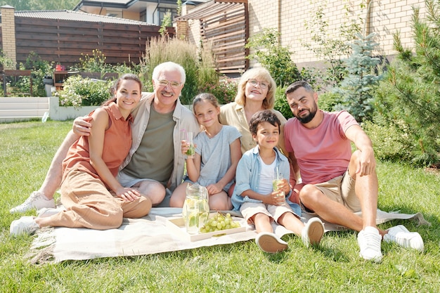 Retrato de uma família moderna de várias gerações fazendo um piquenique no gramado no quintal em um dia ensolarado de verão