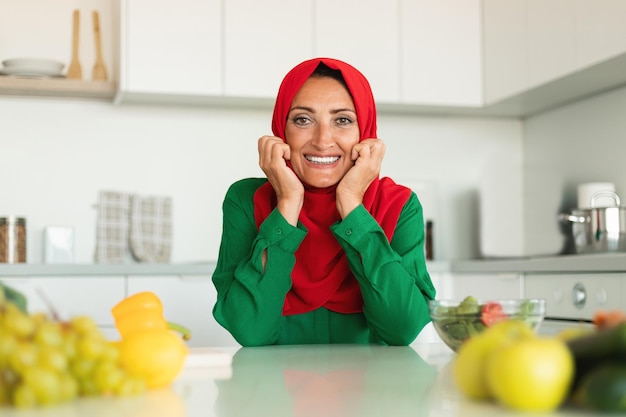 Retrato de uma feliz dona de casa muçulmana de meia-idade posando na cozinha sentada à mesa e sorrindo para o espaço livre da câmera