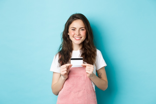 Retrato de uma fofa mulher sorridente, mostrando o cartão de crédito de plástico nas mãos, em pé sobre um fundo azul.