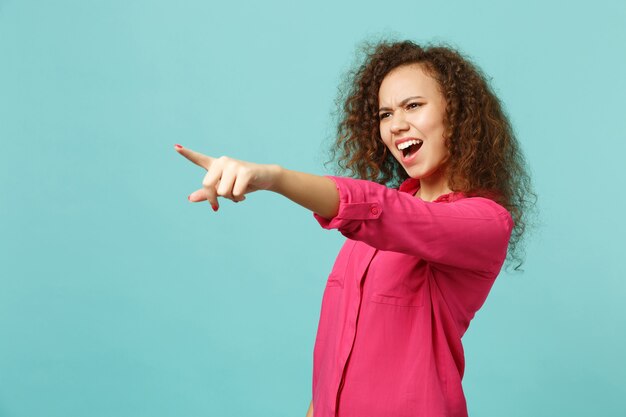 Retrato de uma garota africana perplexa em roupas casuais xingando, apontando o dedo indicador de lado, isolado no fundo da parede azul turquesa. Emoções sinceras de pessoas, conceito de estilo de vida. Simule o espaço da cópia.