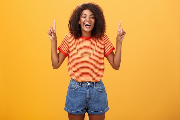 retrato de uma garota afro-americana elegante e feliz, despreocupada, com um penteado afro rindo alto com alegria apontando para cima da parede laranja