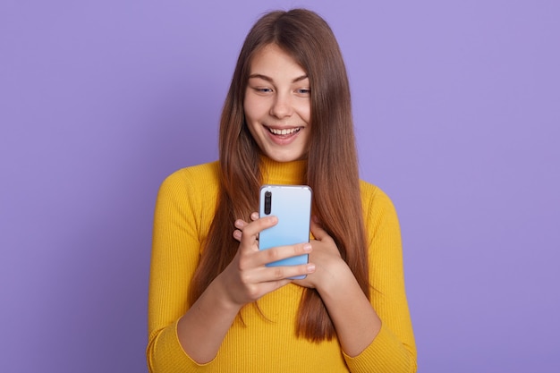 Retrato de uma garota atraente e positiva segurando um telefone inteligente, vestindo camisa amarela, sorrindo com aprovação e satisfação sobre um fundo lilás, olha para o celular