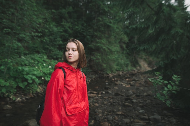 Retrato de uma garota atraente em uma capa de chuva vermelha fica na natureza selvagem da montanha