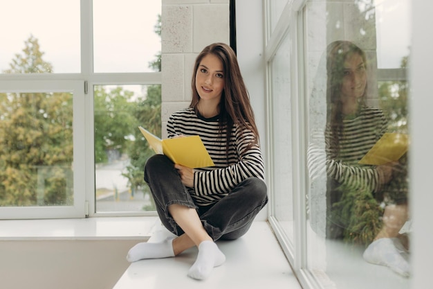 Retrato de uma garota atraente sentada no parapeito da janela e fazendo lição de casa em um notebook