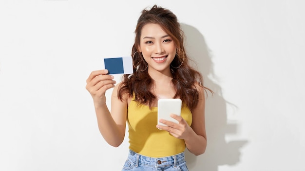 Retrato de uma garota bonita e feliz asiática segurando um smartphone e um cartão de crédito para apresentar compras on-line isoladas sobre um fundo branco
