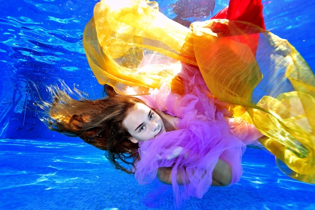 Retrato de uma garota de cabelo comprido que nada e brinca debaixo d'água em uma piscina com tecidos coloridos