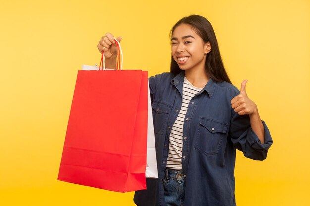 Retrato de uma garota de compras alegre animada em elegante camisa jeans segurando sacos e mostrando os polegares, satisfeito com compras na loja de moda, venda de segunda mão. tiro de estúdio isolado em fundo amarelo