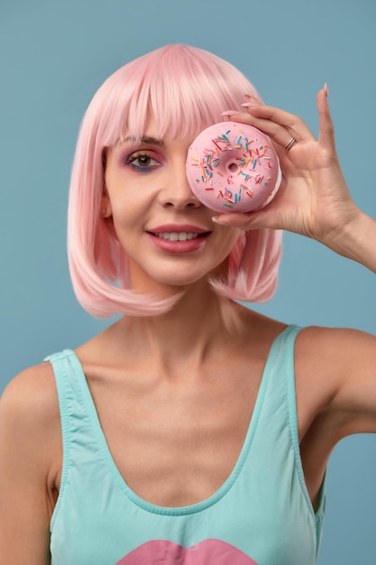 Retrato de uma garota elegante em uma peruca rosa e com uma rosquinha rosa nas mãos mantém o olho mostra a língua