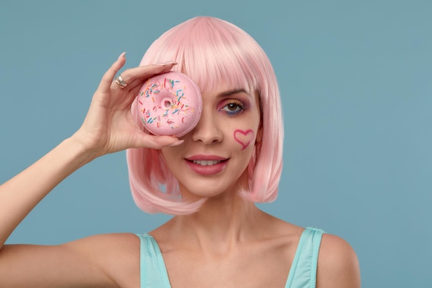 Retrato de uma garota elegante em uma peruca rosa e com uma rosquinha rosa nas mãos mantém o olho mostra a língua