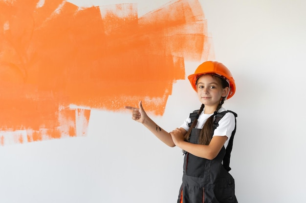 Retrato de uma garota encantadora em um macacão com tinta laranja. no contexto de uma parede com uma mancha. Apontar com o dedo para um espaço vazio para texto.