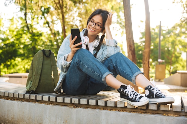 Retrato de uma garota feliz bonito jovem estudante feliz positivo usando óculos, sentado no banco ao ar livre no parque natural, usando o telefone móvel conversando.