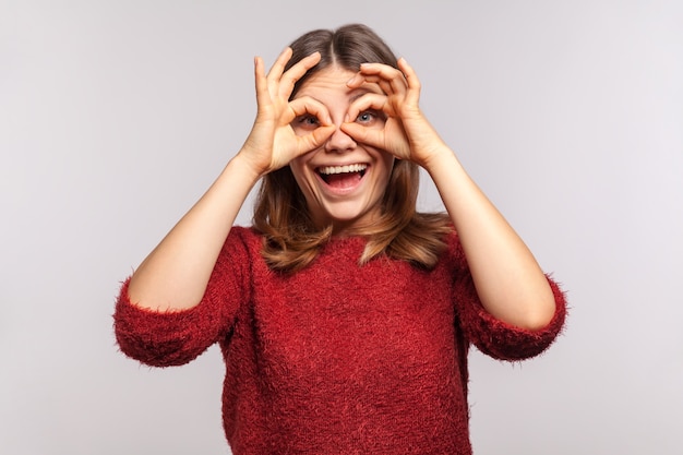 Retrato de uma garota feliz e despreocupada olhando através dos buracos dos dedos, binóculo gesto com a mão