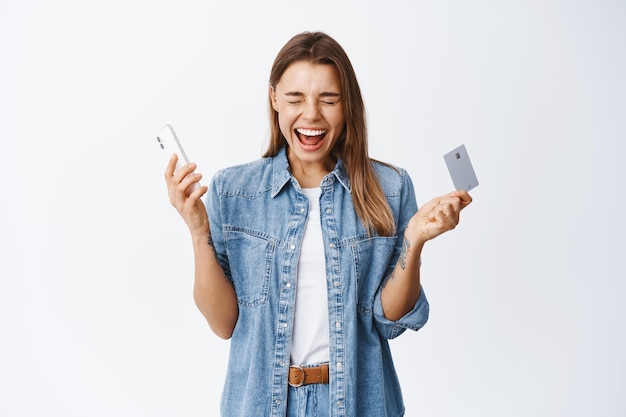 Retrato de uma garota loira animada gritando de felicidade, ganhando dinheiro, segurando um cartão de crédito de plástico e smartphone, parado no branco