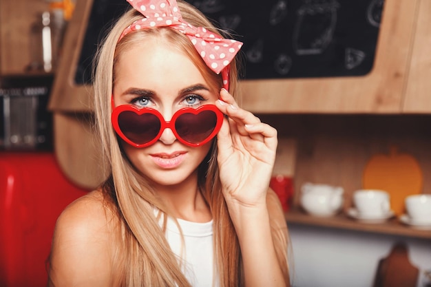 Retrato de uma garota loira sexy de cabelos longos usando óculos de sol olhando para a câmera no loft vermelho confortável cozinha ensolarada