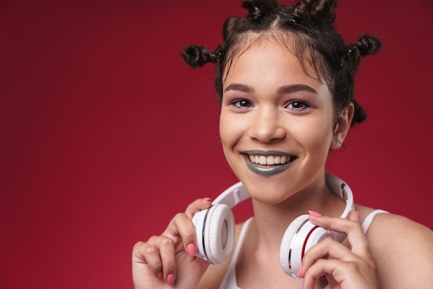 Retrato de uma garota punk alegre com um penteado bizarro e batom escuro sorrindo enquanto ouve música com fones de ouvido isolados sobre a parede vermelha