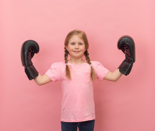 Retrato de uma garotinha alegre e brincalhona em luvas de boxe fazendo gesto de força no fundo rosa