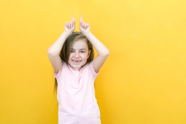 Retrato de uma garotinha alegre e fofa mostrando gesto de chifre de touro piada engraçada de criança atirando dentro de casa fundo amarelo
