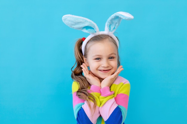 Retrato de uma garotinha com orelhas de coelho em uma jaqueta listrada sorrindo e se entregando O conceito do feriado de Páscoa Lugar de estúdio fotográfico de fundo azul para texto