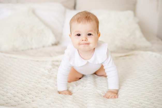 Retrato de uma garotinha de seis meses em uma cama em casa em um quarto brilhante sorrindo um recém-nascido feliz em uma roupa de algodão branco rastejando