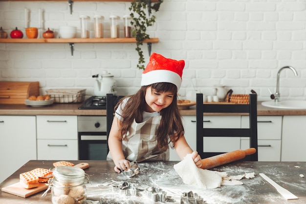 Retrato de uma garotinha feliz em um chapéu de Papai Noel rolando massa na mesa da cozinha uma criança preparando biscoitos de Natal