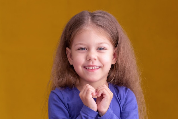 Retrato de uma garotinha sorridente feliz de 5 anos olhando para a câmera segurando a mão em forma de coração em fundo amarelo