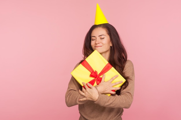 Retrato de uma incrível mulher satisfeita alegre com cone de festa abraçando o melhor presente de feriado e fechando os olhos, sorrindo com expressão satisfeita, comemorando aniversário, aniversário. estúdio tiro fundo rosa