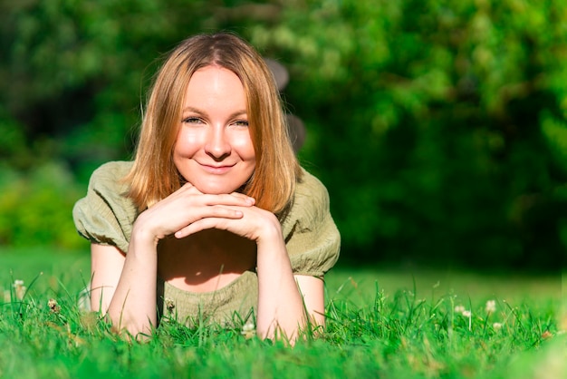 Retrato de uma jovem adolescente feliz alegre positiva, jovem muito alegre está deitada de bruços na grama verde, sorrindo, olhando para a câmera, aproveitando o bom dia de verão. Copie o espaço, coloque para o texto