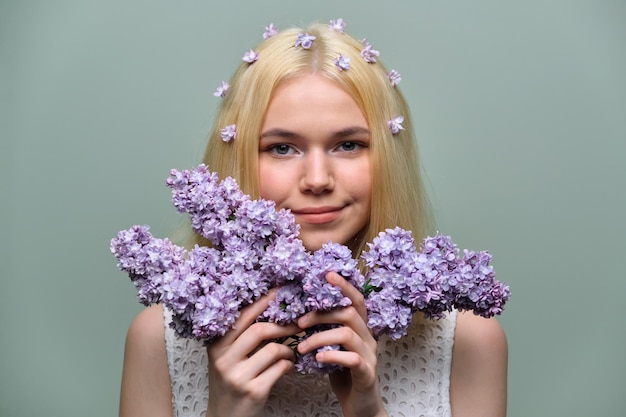 Retrato de uma jovem adolescente loira com flores lilás violetas no cabelo e nas mãos, fundo verde pastel do estúdio. Pele saudável e maquiagem natural para adolescentes