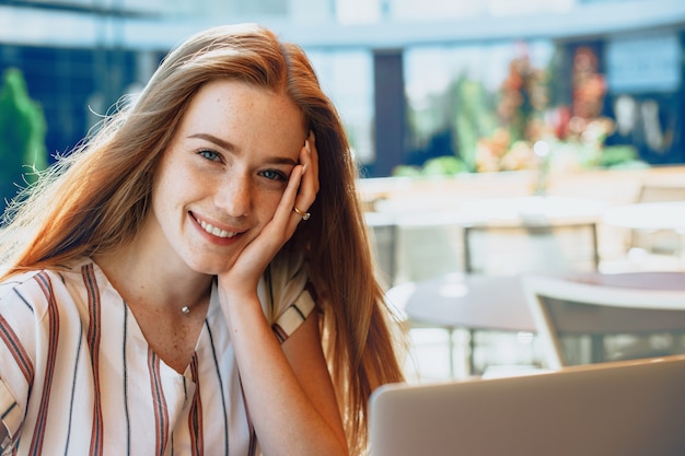 Retrato de uma jovem adorável com cabelo ruivo e sardas sorrindo enquanto toca o rosto com uma mão que trabalha em um laptop ao ar livre em uma cafeteria.