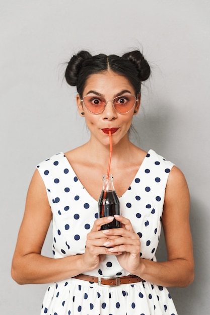 Foto retrato de uma jovem adorável com vestido de verão e óculos de sol isolados, segurando uma garrafa com refrigerante