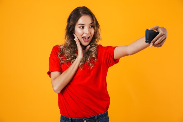 Retrato de uma jovem adorável e alegre em pé isolado na parede amarela, tirando uma selfie