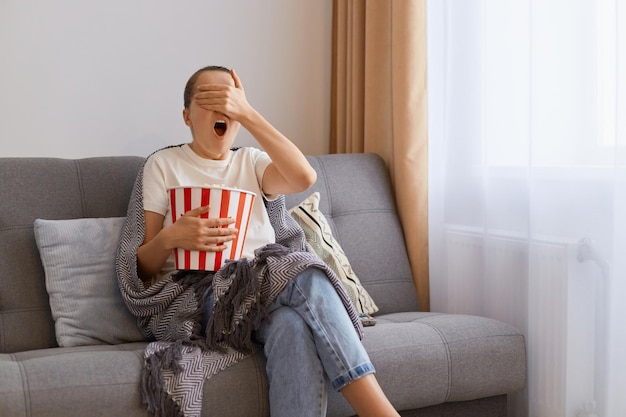 Retrato de uma jovem adulta assustada vestindo camiseta branca sentada no sofá com pipoca e controle remoto assistindo filme de terror cobrindo os olhos com palma vê momento de medo