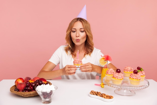 Retrato de uma jovem adulta segurando o bolo de aniversário e soprando vela, celebração sozinha, fazendo desejo, fêmea em t-shirt casual e cone de festa. Tiro de estúdio interior isolado no fundo rosa.