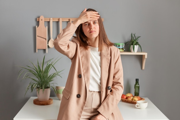 Retrato de uma jovem adulta triste e doente vestindo terno bege em pé perto da mesa na cozinha em casa mantém a mão na testa sofrendo de dor de cabeça com alta temperatura