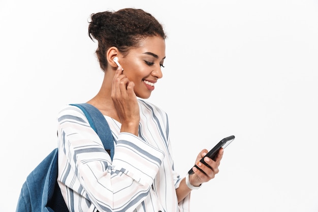 Retrato de uma jovem africana atraente carregando uma mochila em pé, isolada na parede branca, ouvindo música com fones de ouvido sem fio, segurando um telefone celular