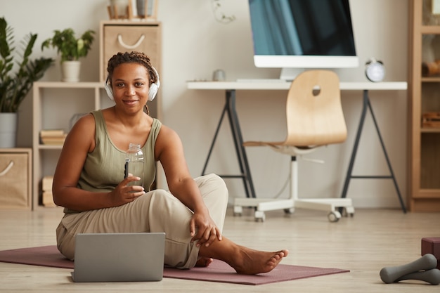 Retrato de uma jovem africana em fones de ouvido, bebendo água e enquanto está sentado na esteira de exercícios e descansando após o treino