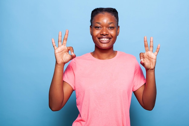 Retrato de uma jovem afro-americana alegre positiva sorrindo para a câmera mostrando sinal de ok em pé no estúdio com fundo azul. Adolescente otimista aprova a ideia. Sinal de acordo