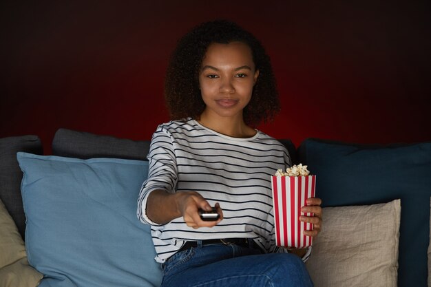 Foto retrato de uma jovem afro-americana assistindo tv em casa e segurando pipoca enquanto está sentada no sofá em um quarto escuro