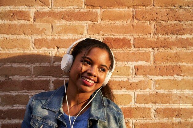 Retrato de uma jovem afro-americana com fones de ouvido, ouvindo música e sorrindo engraçado em uma parede de tijolos