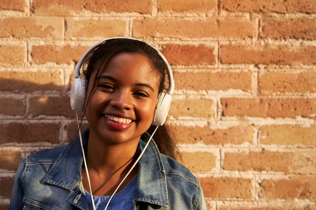 Retrato de uma jovem afro-americana com fones de ouvido, ouvindo música e sorrindo engraçado em uma parede de tijolos