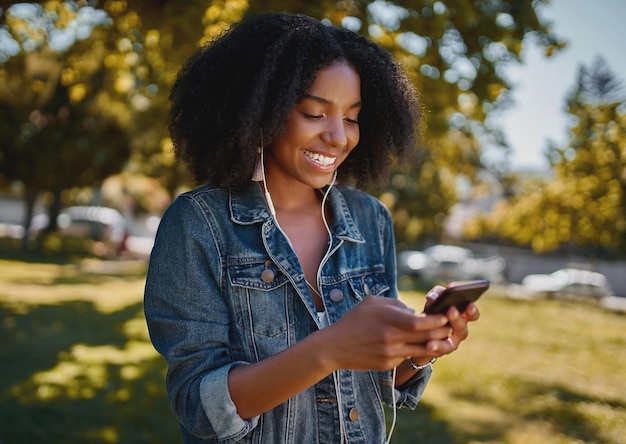 Retrato de uma jovem afro-americana de moda sorridente enviando mensagens de texto no telefone celular de pé no parque ouvindo a marca