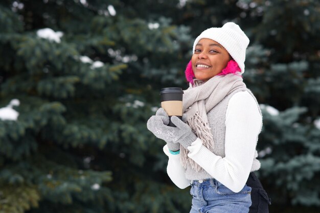 Retrato de uma jovem afro-americana feliz em um dia de neve de inverno no parque de neve, sorrindo, bebendo café