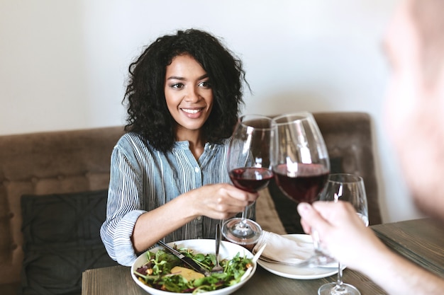 Retrato de uma jovem afro-americana no restaurante com um copo de vinho tinto na mão e salada na mesa. Menina muito sorridente com cabelo escuro e encaracolado sentada em um café e bebendo vinho com um amigo