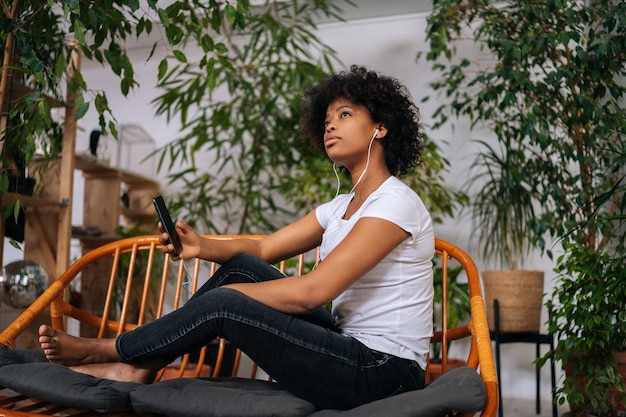 Retrato de uma jovem afro-americana pensativa ouvindo música do celular pensando olhando para longe sentado no sofá em casa