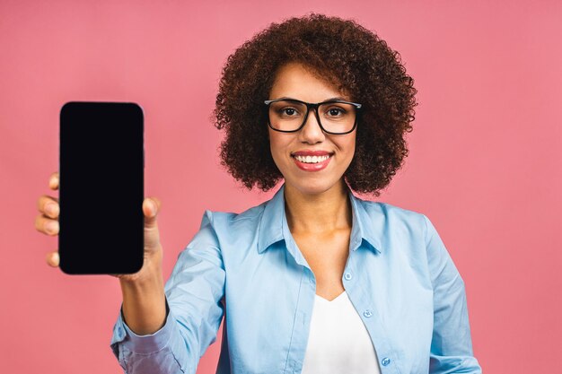 Retrato de uma jovem afro-americana sorridente segurando o celular de tela em branco e mostrando o dedo isolado sobre o fundo rosa