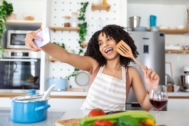 Retrato de uma jovem afro-americana sorridente tomando selfie com um smartphone enquanto cozinha na cozinha em casa