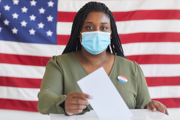 Retrato de uma jovem afro-americana usando máscara, colocando o boletim de voto nas urnas e, em pé contra a bandeira americana no dia da eleição, copie o espaço