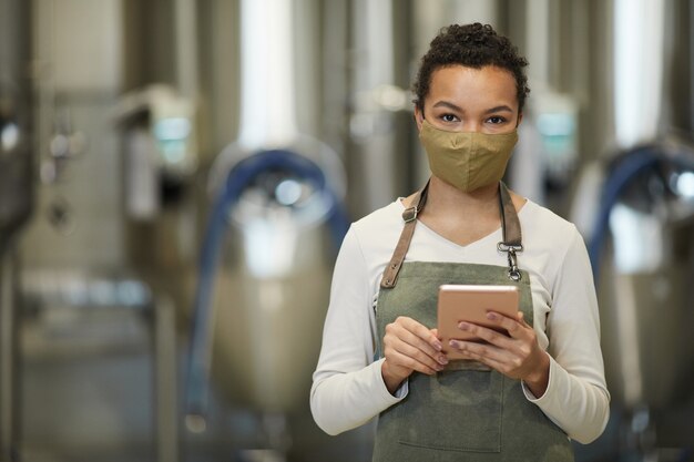 Foto retrato de uma jovem afro-americana usando máscara e avental enquanto posava na oficina de uma cervejaria industrial moderna, copie o espaço da cintura para cima