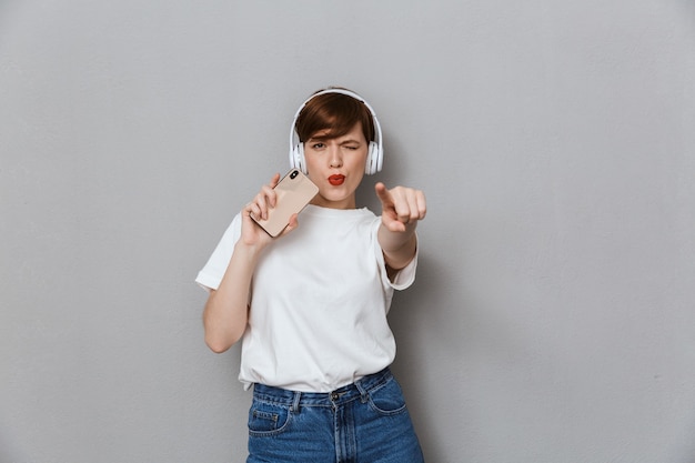 Retrato de uma jovem alegre cantando enquanto ouve música com fones de ouvido e smartphone isolados sobre uma parede cinza