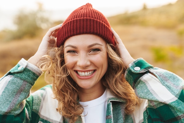 Retrato de uma jovem alegre, caucasiana, usando chapéu e camisa xadrez, sorrindo enquanto caminha ao ar livre